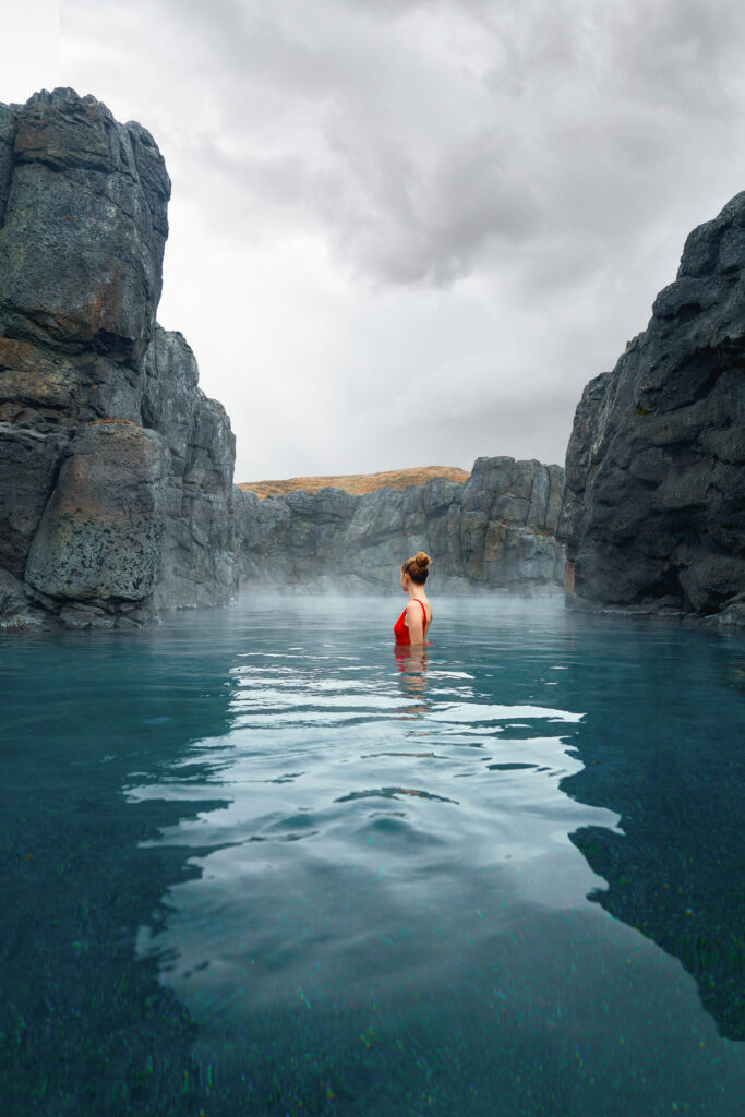 A woman in a red swimsuit stands centered in the calm, steamy waters of the Sky Lagoon in Iceland, flanked by rugged black cliffs under a moody sky in Iceland