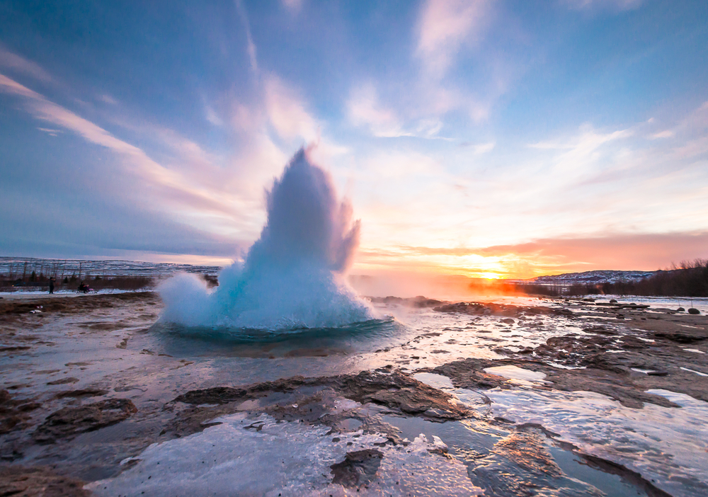 Geysir eruption in Iceland at sunset, with steam and hot water shooting into the air against a pastel sky.





