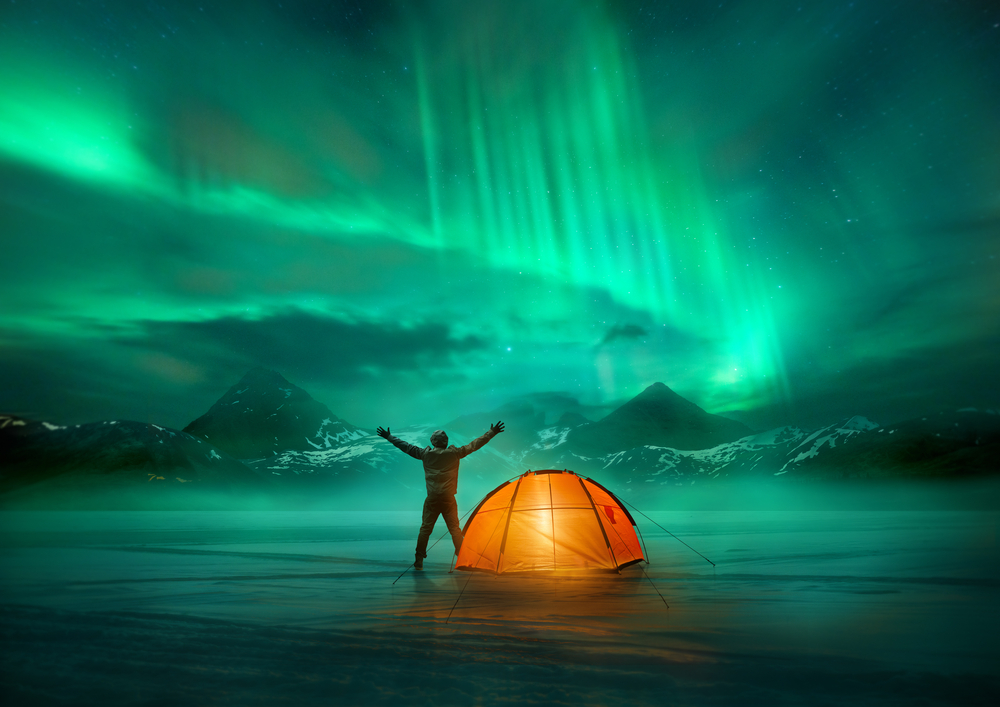 A figure celebrates under the ethereal glow of the Northern Lights in Iceland, with a bright orange tent providing a stark contrast against the icy wilderness