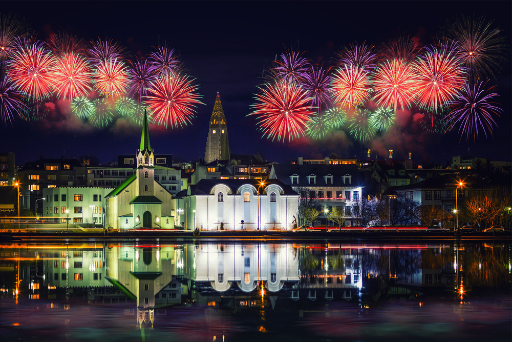 Vibrant fireworks light up the night sky above the iconic Reykjavik skyline, with reflections shimmering in the calm waters below