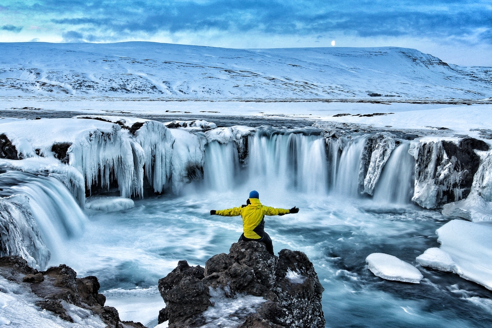 A lone figure stands with arms outstretched before the powerful and partially frozen Godafoss Waterfall in Iceland, under a pale winter sky.