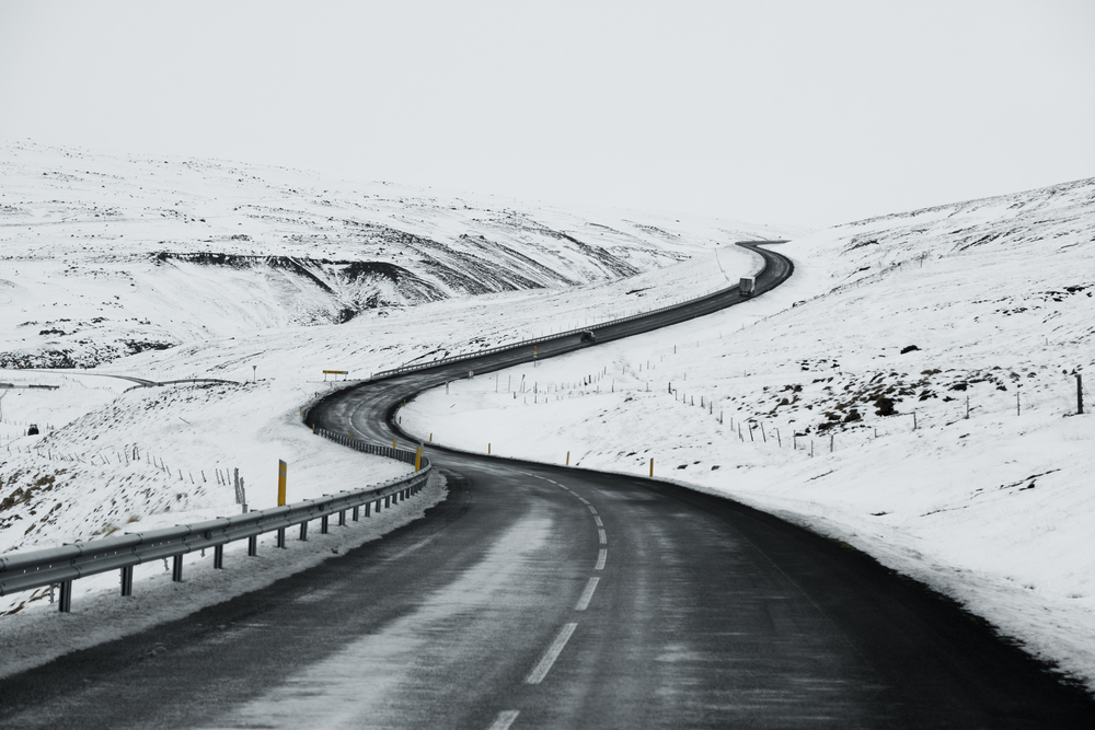 A curving icy road cuts through a snowy landscape in Iceland, with stark white fields meeting a grey overcast sky.