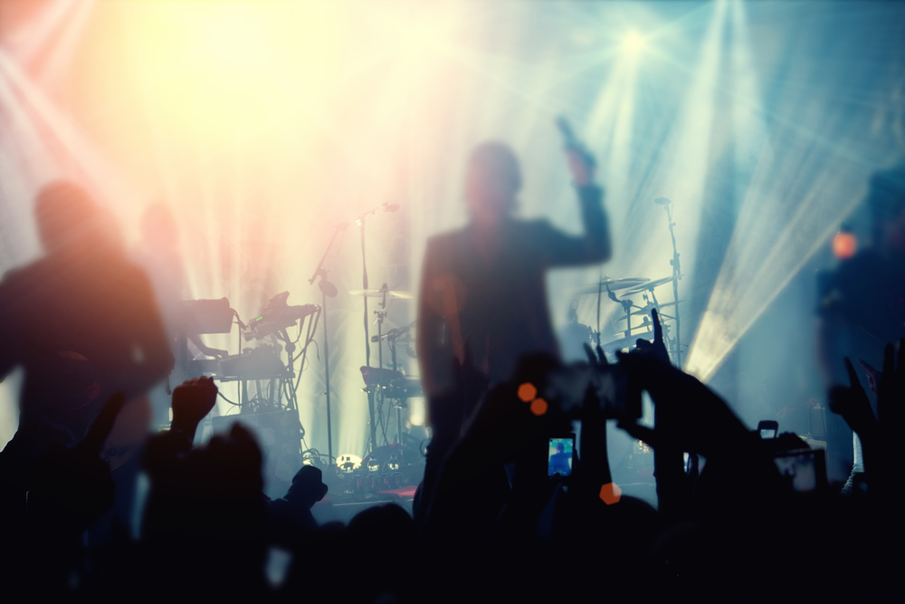 Blurred silhouette of a singer on stage at a music festival, with hands of the audience raised and stage lights creating a bright, vibrant halo effect