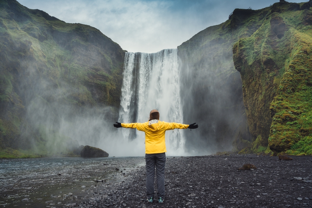 A person in a bright yellow jacket stands with arms outstretched facing skogafoss amid misty, green, moss-covered cliffs, embodying a sense of freedom and awe in nature.