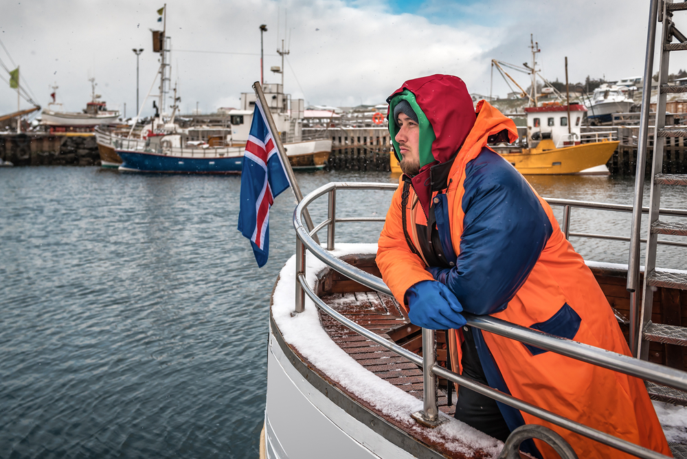 A person in a colorful orange and blue hooded jacket standing on a boat, leaning on the railing with a contemplative expression, overlooking a harbor with snowflakes falling and fishing boats docked in the background, with the Icelandic flag prominently displayed in the foreground.
