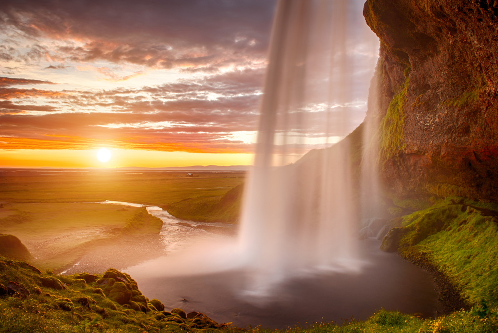 Seljalandsfoss waterfall in Iceland during sunset, with the sun casting a warm glow over the green landscape and cascading water