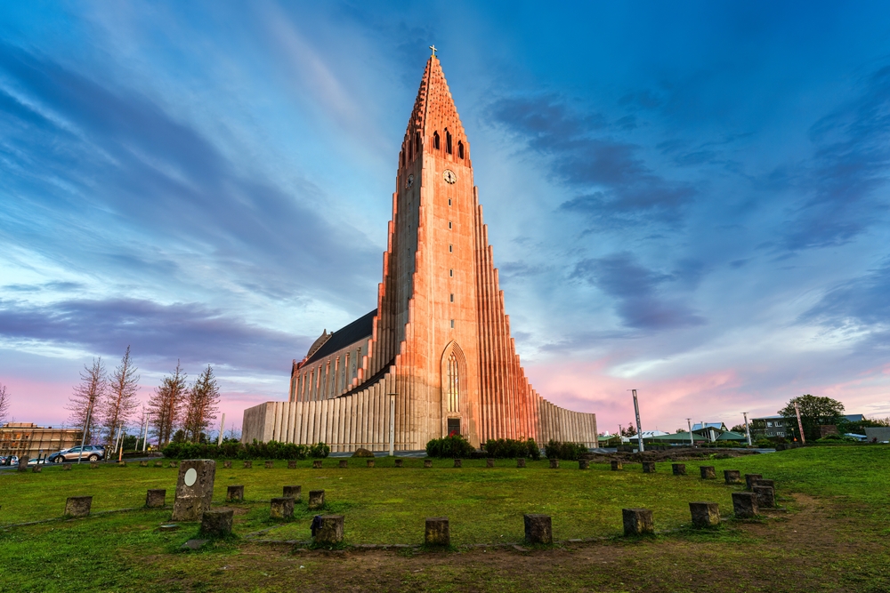 The Hallgrimskirkja church in Reykjavik, Iceland, is bathed in the warm light of the setting sun, with a clear blue sky above and small wooden stumps dotting the foreground