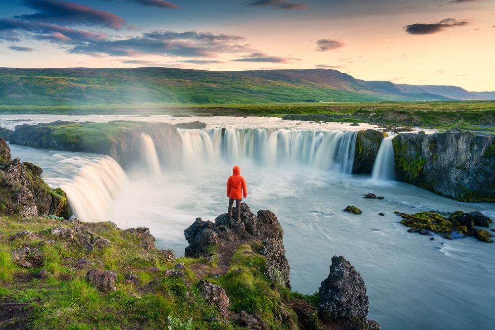 A solitary figure in a red jacket stands on a rocky outcrop, gazing at the majestic Godafoss Waterfall under the soft glow of a sunset in Iceland