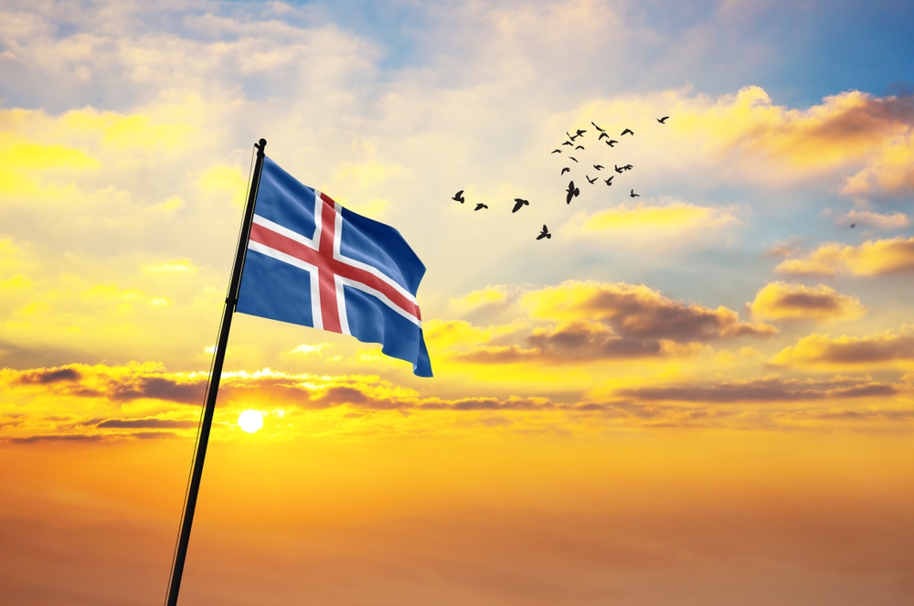 Icelandic flag fluttering proudly against a serene sunset sky, with a flock of birds flying in the background.
