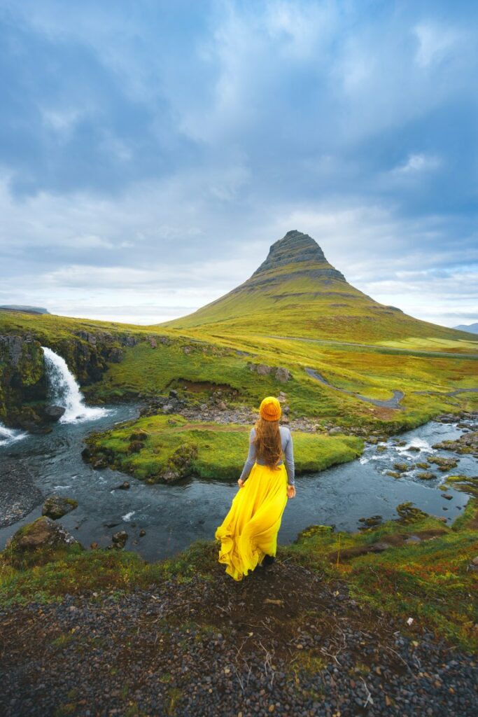 Woman in a flowing yellow skirt overlooking Kirkjufellfoss and the Kirkjufell mountain on a cloudy day.