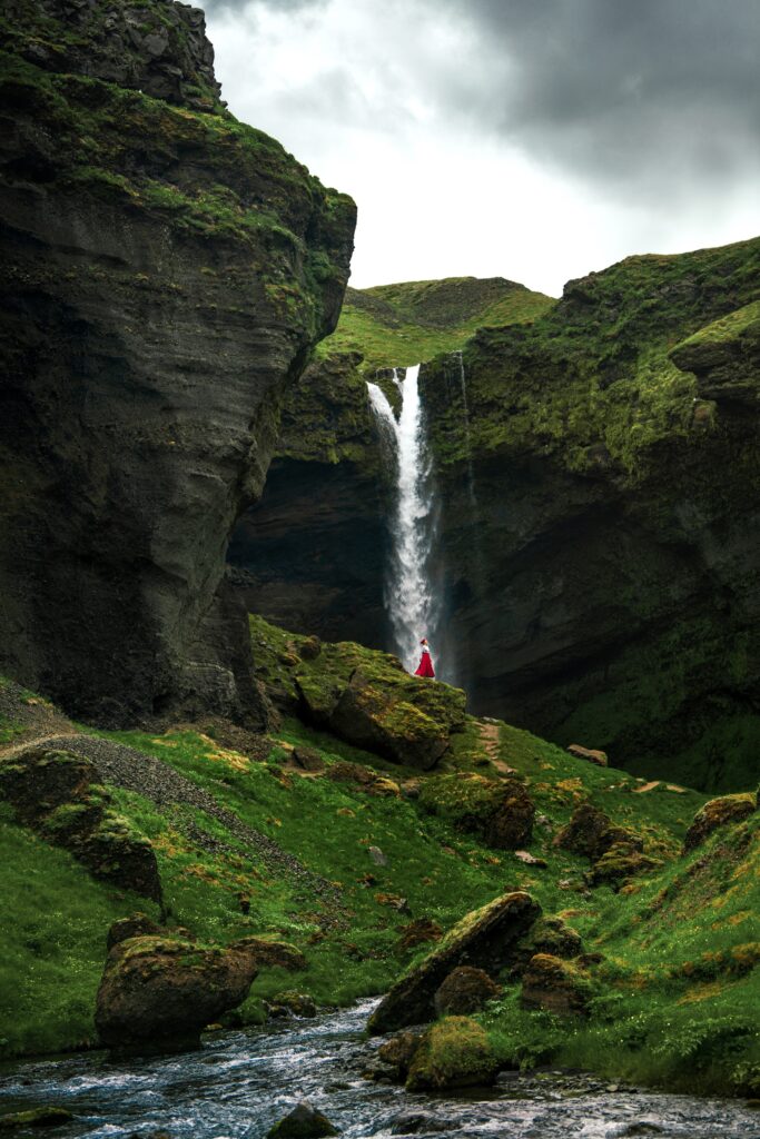 Small figure of a woman in a red skirt standing in front of the Kvernufoss Waterfall in a mossy gorge.