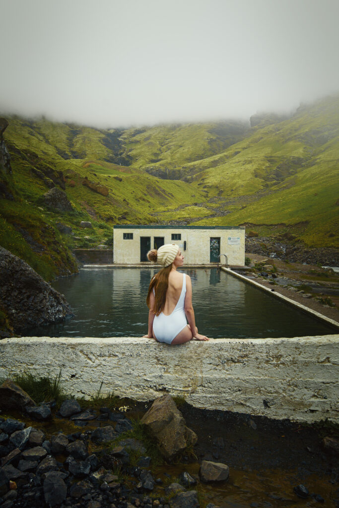Low clouds over the Seljavallalaug Hot Spring with a woman wearing a swimsuit and beanie hat.