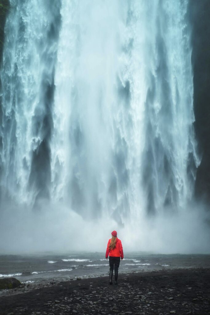 Woman in a red coat and hat standing at the bottom of the massive Skogafoss Waterfall.