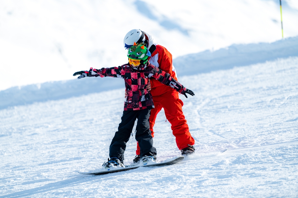 Adult snowboard instructor guiding a young child on a snowboard, both wearing colorful winter gear and safety helmets against a snowy Iceland mountain backdrop.




