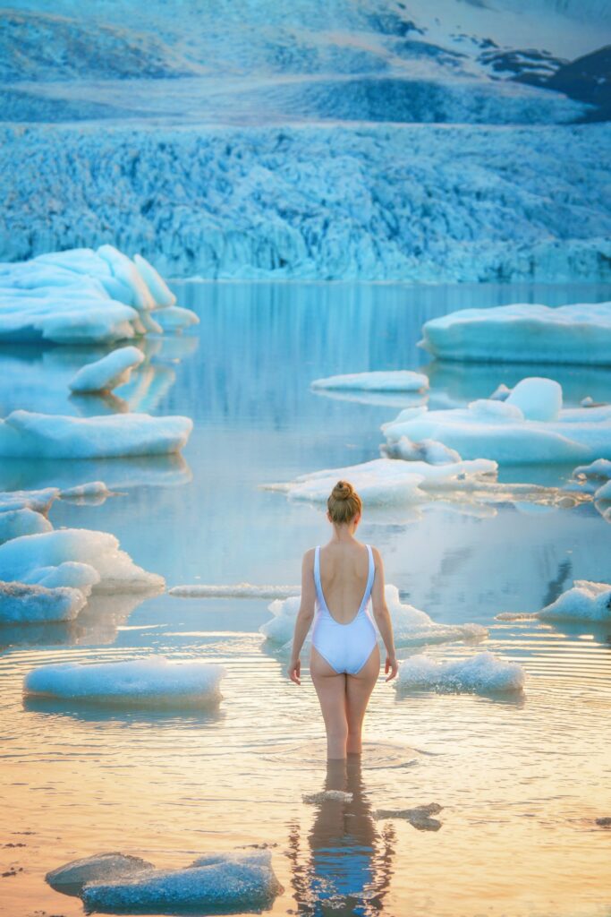 Woman in a white one piece swimsuit standing ankle deep in the glacier lagoon with icebergs.