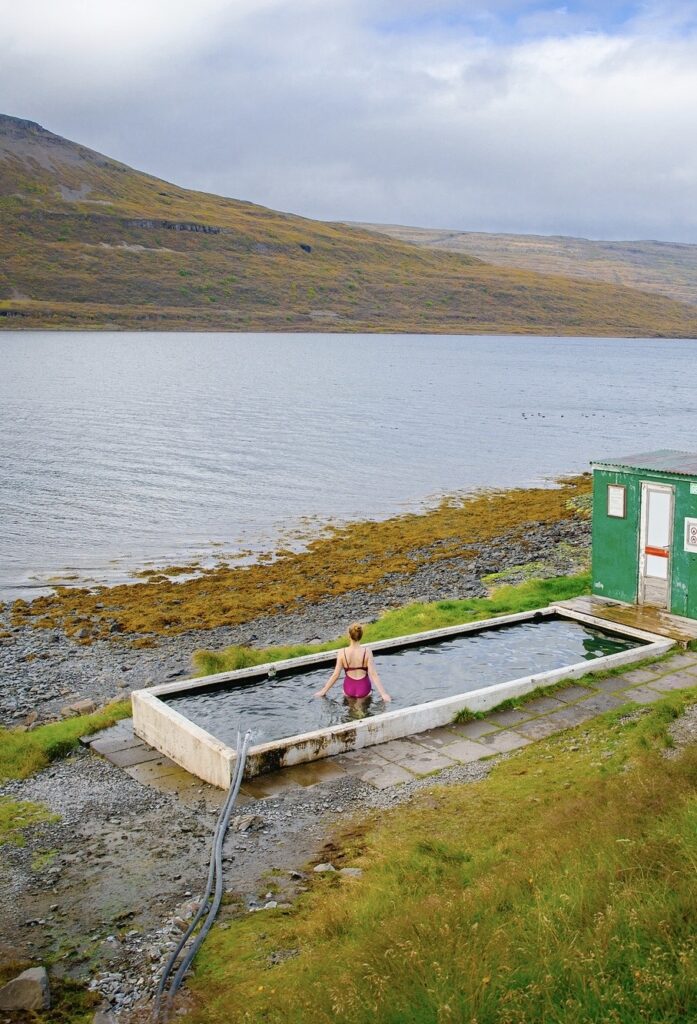 standing in the water at the rectangular man-made hot swimming pool Hörgshlíðarlaug looking at the ocean with a green changing hut to the right