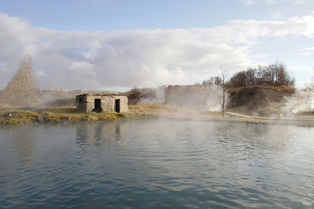 Secret Lagoon in Iceland, showcasing a tranquil lake with steam rising from the water, a small hill in the background, and a rustic stone structure on the shore, under a clear blue sky with light clouds."