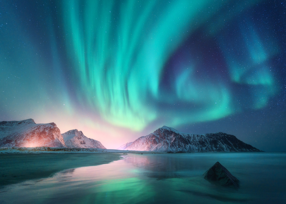 The Northern Lights swirl in a mesmerizing dance of green above a quiet, snow-dusted beach, framed by darkened hills