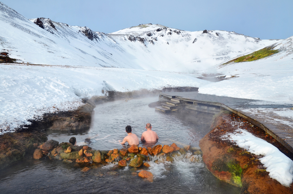Two people enjoying a geothermal hot spring surrounded by snow-capped mountains in Iceland, capturing the unique experience of Icelandic winters in March.