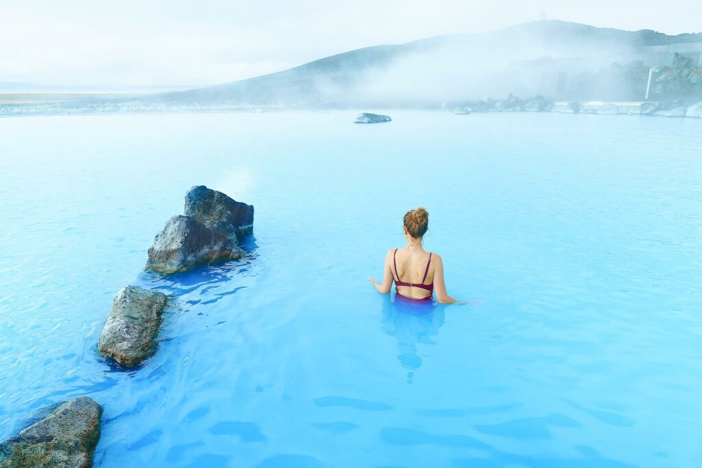standing in the blue waters of the Myvatn Nature Baths with mountains in the distance and a line of rocks in the water to the left