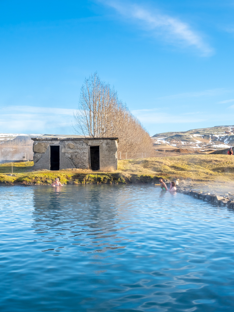 3 bathers in the Secret Lagoon pool on a sunny winter day with a small hut in the background and bare trees and snowy mountains behind that