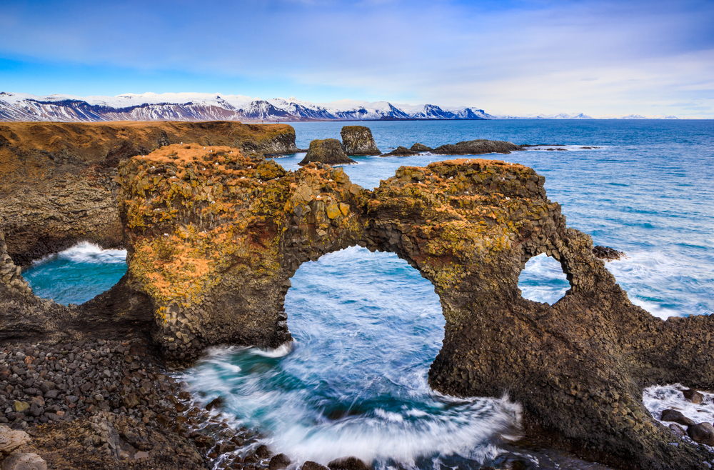 Rugged sea arch along the coast with mountains in the background.