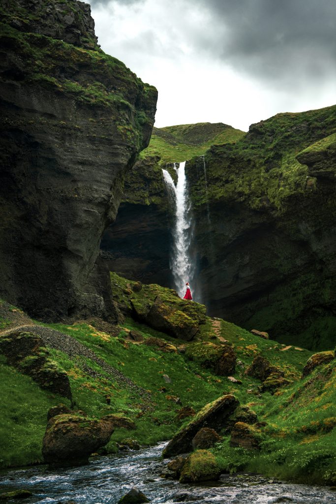 Woman in a red skirt standing an a mossy, green, and rugged hill under the Kvernufoss Waterfall on a cloudy day.