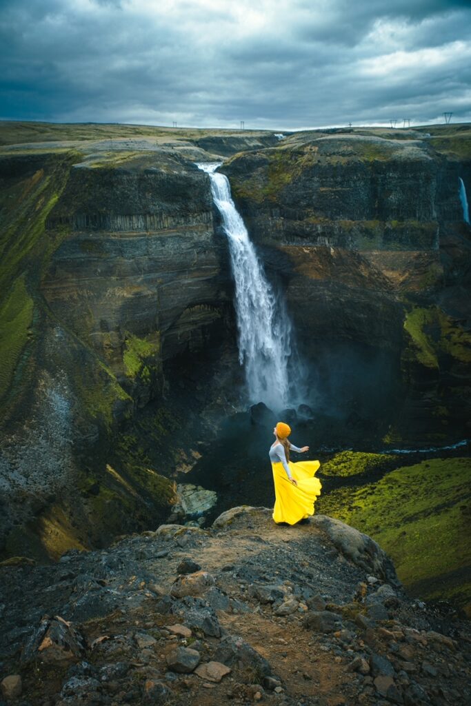 standing on the edge of a canyon in a yellow skirt across from Haifoss Waterfall