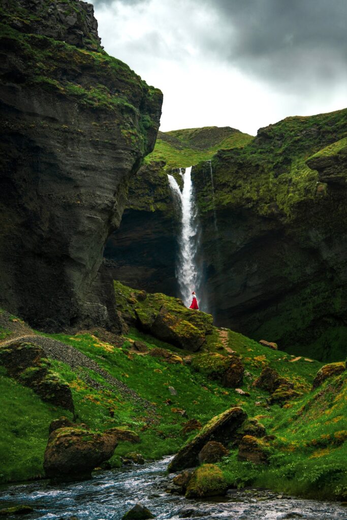 a zoomed out view of a woman standing at the base of Kvernufoss Waterfall in a red skirt surrounded by mossy landscape