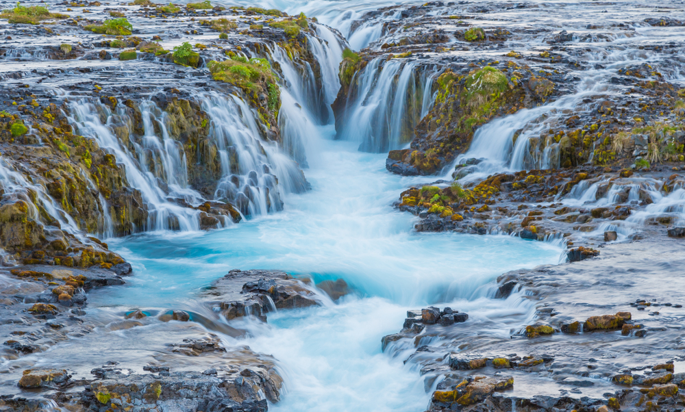 a close up view of the many, very blue cascades of Bruarfoss Waterfall flowing over lava rock and moss