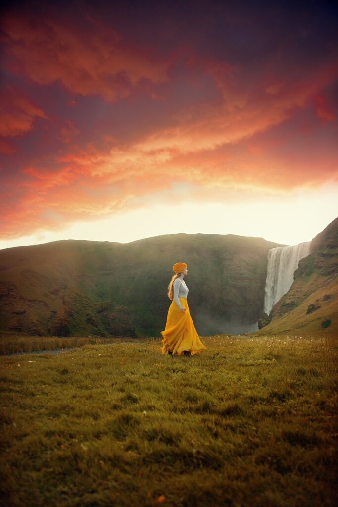 a woman in a yellow skirt walking on grass with a glimpse of Skogafoss Waterfall, one of the best waterfalls near Reykjavik, in the distance