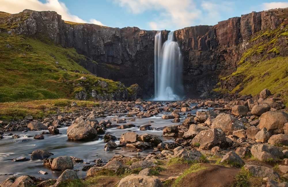 a slightly pulled back view of Gufufoss flowing over the cliff with mossy slopes on either side of it and a rocky river in the foreground