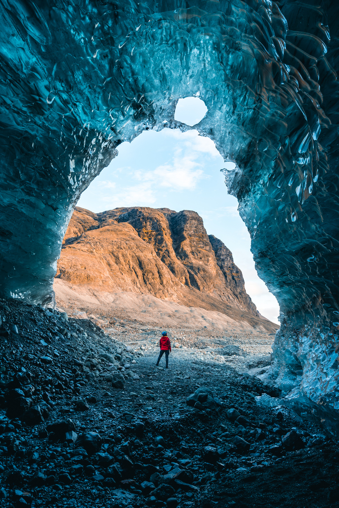 A person in a red jacket stands at the entrance of a massive ice cave, framed by towering, translucent blue ice walls. They face outward towards a rugged, sunlit mountain landscape, creating a dramatic contrast between the icy interior of the cave and the warm, earthy tones of the mountains beyond. The scene captures the awe-inspiring scale and natural beauty of the environment.