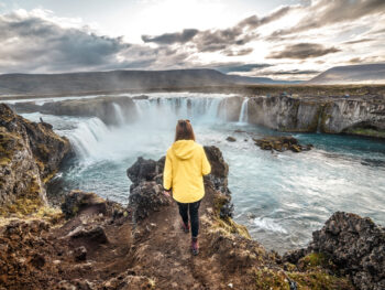 woman standing on the edge of a waterfall in Iceland wearing a yellow jacket and black pants illustrating what to wear in Iceland in december