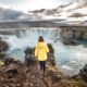 woman standing on the edge of a waterfall in Iceland wearing a yellow jacket and black pants illustrating what to wear in Iceland in december