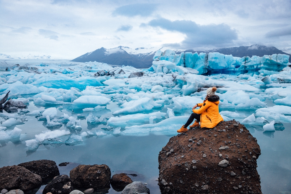 A person dressed in a bright yellow jacket sits on a large rock, gazing at a serene icy landscape with floating icebergs and distant snow-capped mountains.