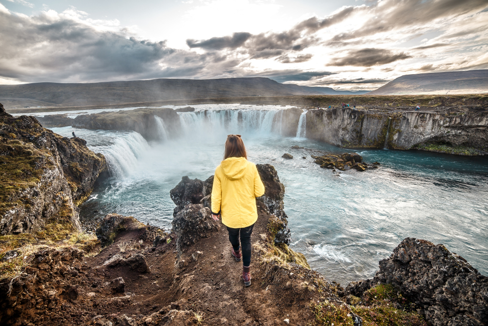 woman standing on the edge of a waterfall in Iceland wearing a yellow jacket and black pants illustrating what to wear in Iceland in december