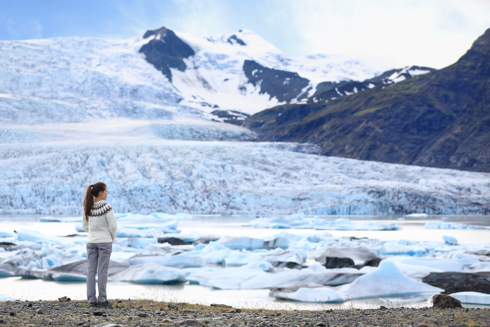 a woman in a white Icelandic sweater and grey pants standing at the edge of a glacier lagoon