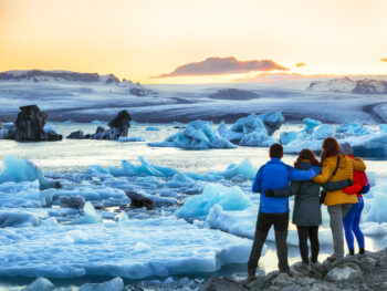 a group of tourists with their arms around each other facing away from the camera wearing winter clothes and looking at a glacier lagoon