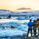 a group of tourists with their arms around each other facing away from the camera wearing winter clothes and looking at a glacier lagoon