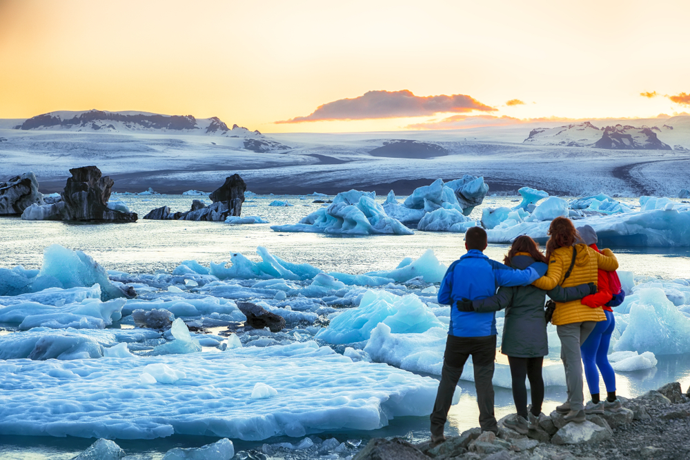 a group of tourists with their arms around each other facing away from the camera wearing winter clothes and looking at a glacier lagoon