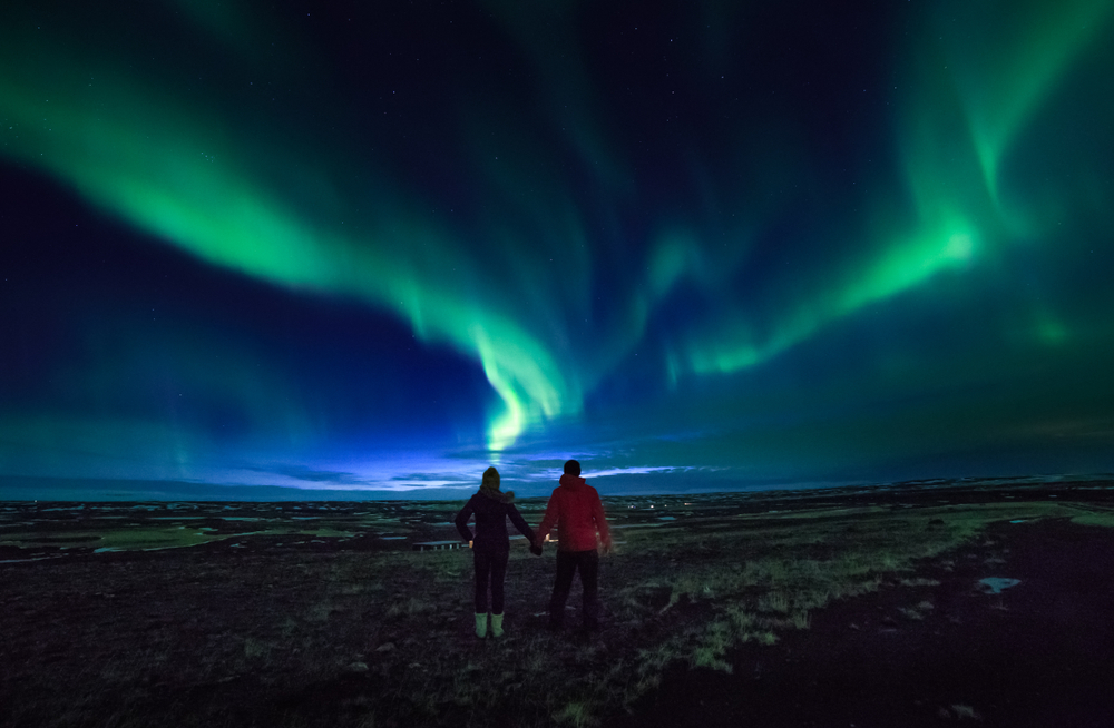 a couple holding hands wearing winter gear watching the Northern Lights dance across the sky