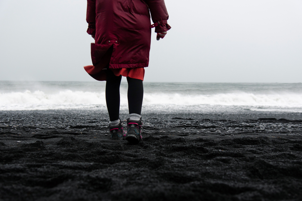  the waist down of a person in a red coat walking away from the camera towards the ocean wearing hiking boots on the Reynisfjara black sand beach