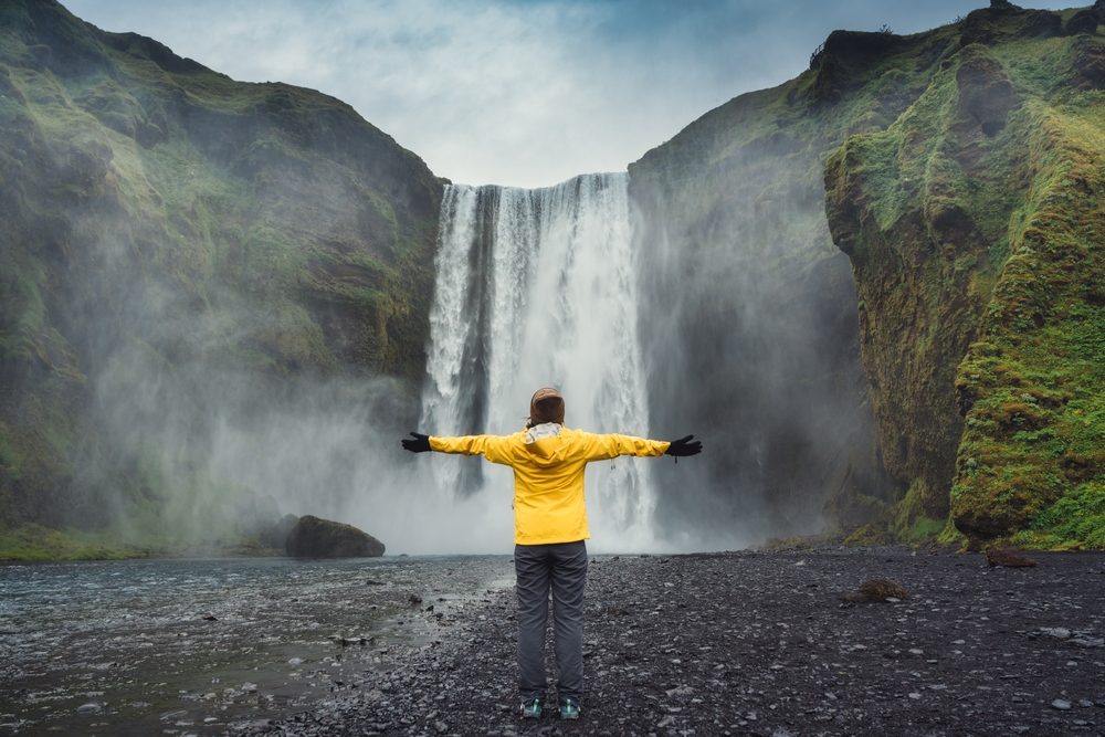 a woman in a yellow rain jacket, grey rain pants, a brown beanie, and black gloves facing away from the camera with her arms outstretched looking at Skogafoss waterfall