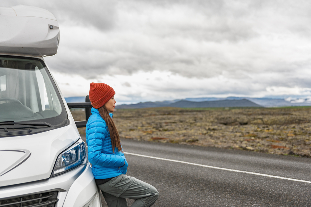 A woman knows what to wear to Iceland in August when traveling: her puffer jacket, beanie and cargo pants are comfortable as she leans against a travel van.