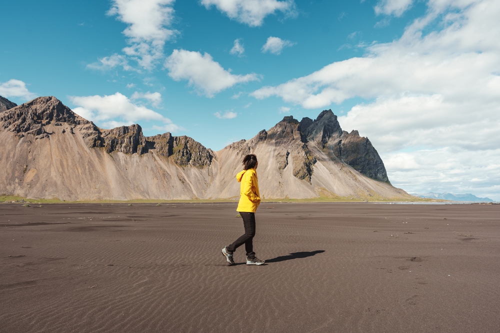 A woman walks along the coast, mountains and dunes behind her, in black pants, a yellow wind breaker, and hiking shoes.