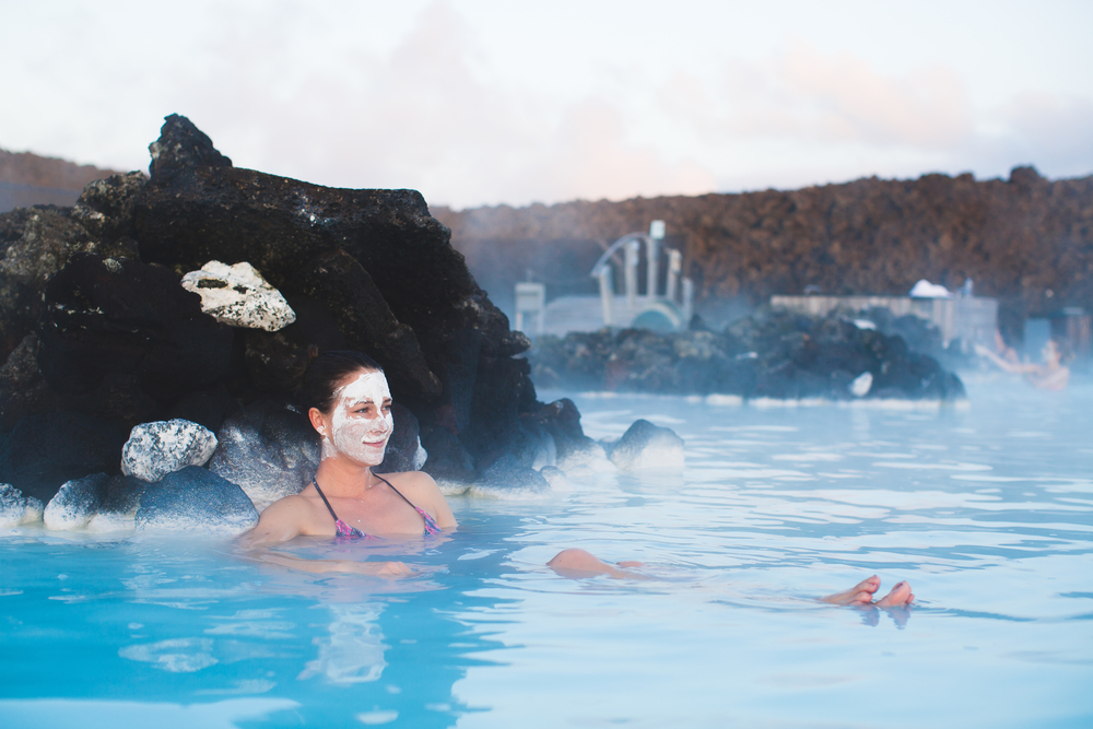 A woman sits in the blue lagoon in a bikini with mud on her face, relaxing in the steam.