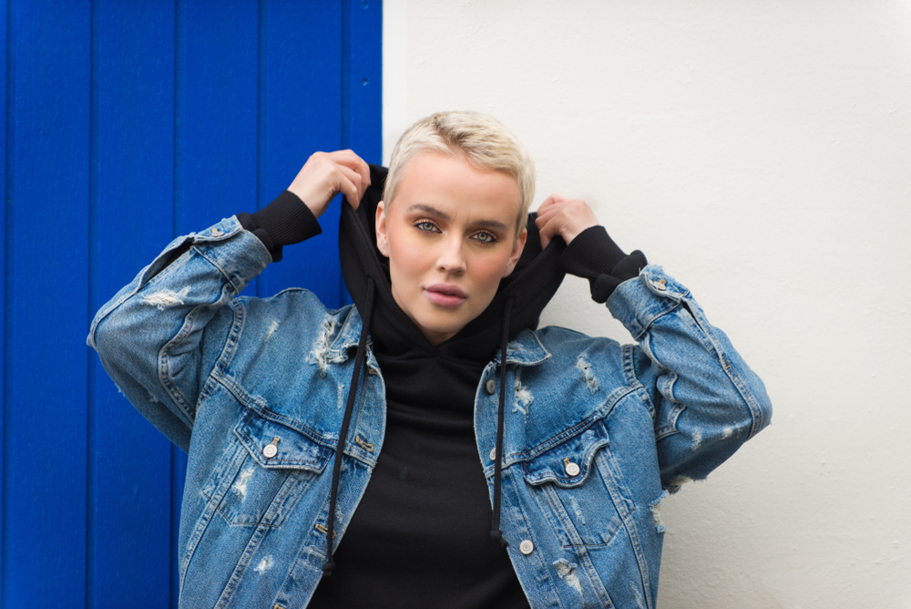 A model stands against a blue and white wall in a city in Iceland wearing a black hoodie with a jean jacket over it to add style to warmth.