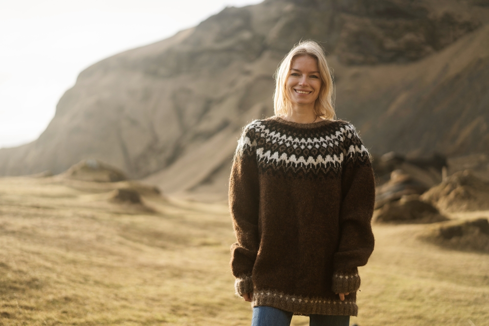 A young blonde girls stands in the green landscapes of Iceland, knowing what to wear to Iceland in July, as she is comfortable in her wool, oversized sweater and jeans.