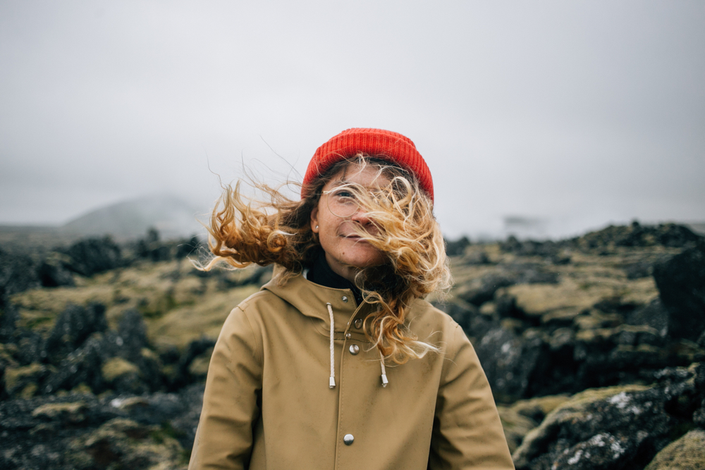 A young woman stands on a hike in Iceland, pausing in the greenery as the wind blows her hair wild as she wears a windbreaker and a red hat and glasses.
